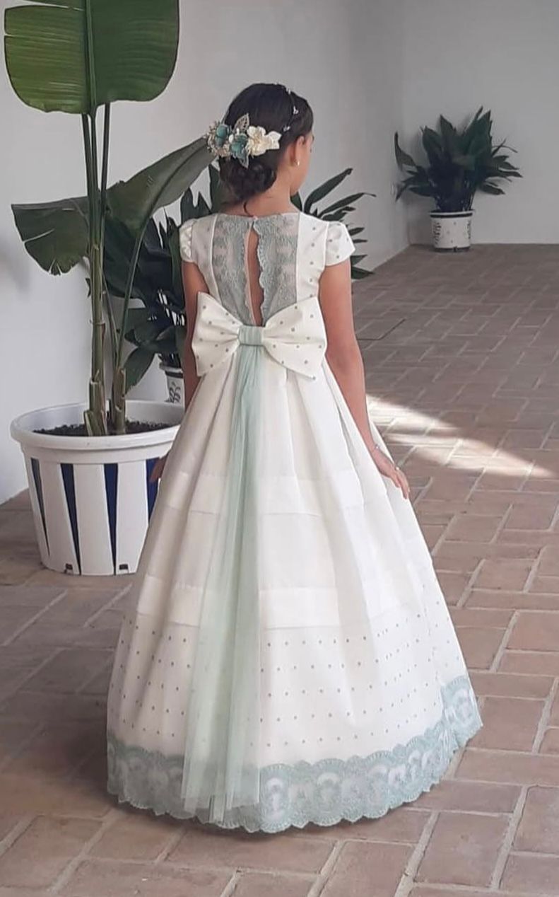 Niña con vestido blanco con lazo, espalda abierta, encaje y tocado floral.