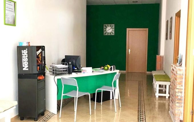 Pequeña sala de estar de oficina con pared decorativa verde, escritorio, sillas, máquina expendedora y pasillo.