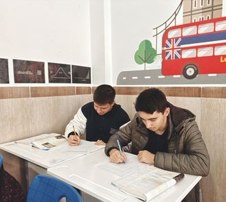Dos estudiantes escribiendo en una mesa en un aula luminosa con murales de temática londinense.