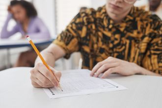 Una persona escribe en un examen con un lápiz en un aula, con otro estudiante borroso al fondo.