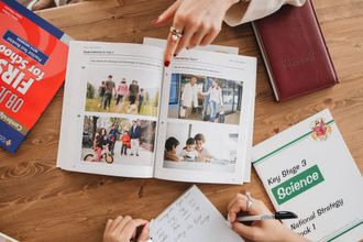 Manos y cuadernos rodean un libro de fotos abierto sobre un escritorio, con un libro de texto de ciencias cerca.