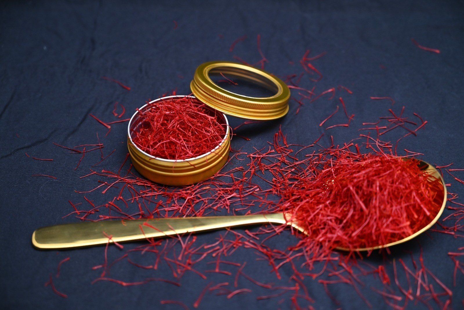 Rote Safranfäden füllen eine goldene Dose und einen passenden Löffel, die auf einem dunkelblauen Hintergrund verstreut sind.