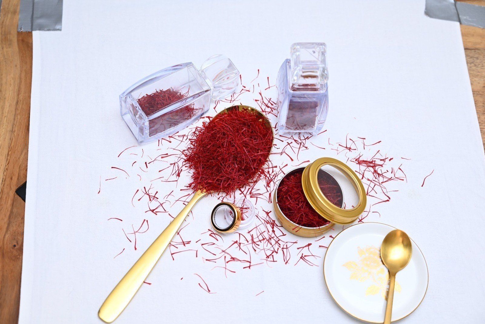 Ein goldener Löffel, eine kleine goldene Dose und Glasbehälter, gefüllt mit tiefroten Safranfäden auf weissem Grund.