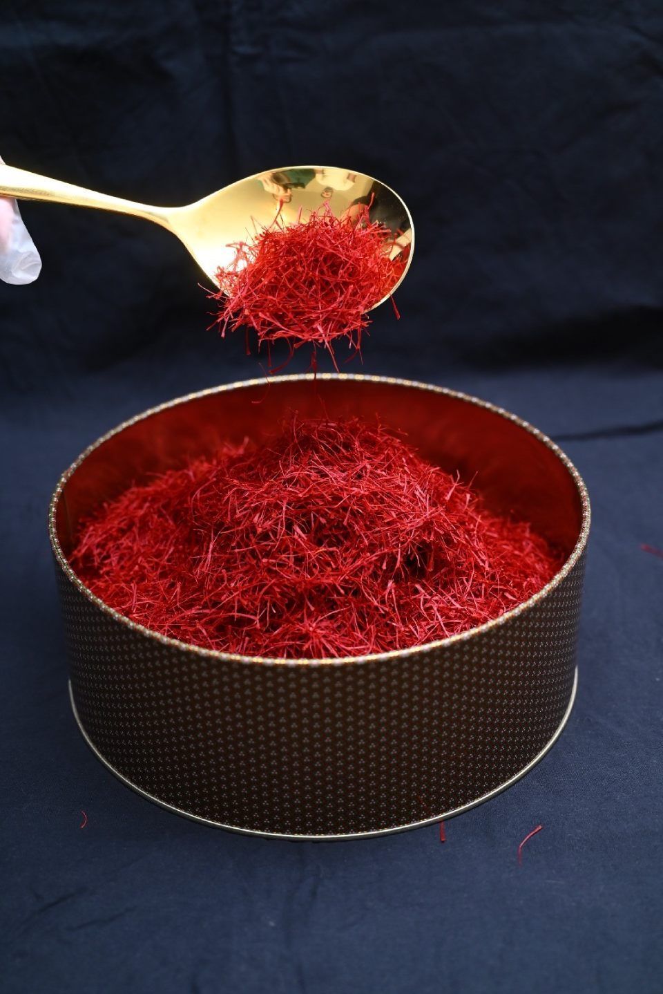 Ein goldener Löffel hält leuchtend rote Safranfäden über einem runden, strukturierten braunen Behälter, der mit weiterem Safran gefüllt ist.