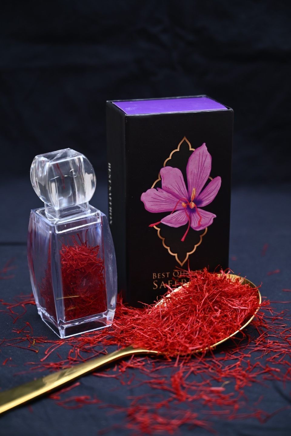 Neben einer schwarzen Schachtel und einem goldenen Löffel, der überquillt vor Safran, steht ein durchsichtiges Glasgefäß, gefüllt mit roten Safranfäden.