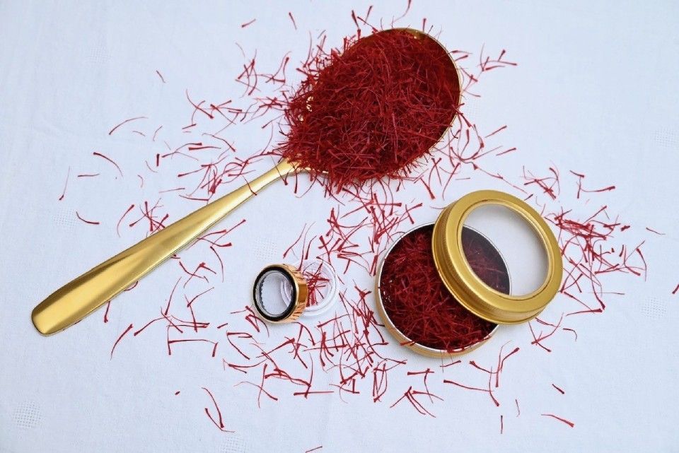 Ein goldener Löffel, bedeckt mit tiefroten Safranfäden, liegt neben einem kleinen, offenen Metallbehälter, der mit dem Gewürz gefüllt ist.