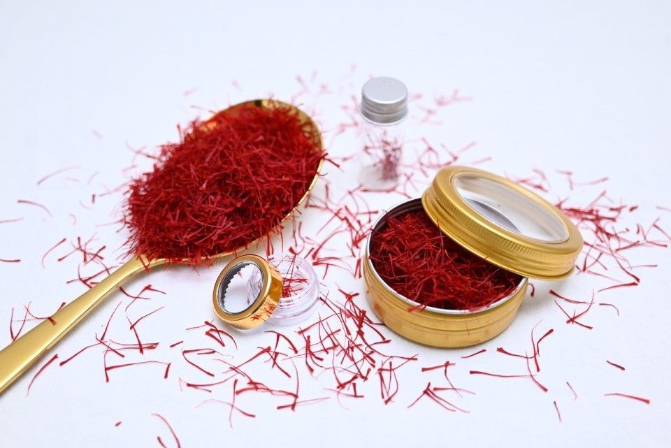 Ein Löffel voll leuchtend roter Safranfäden neben einer kleinen, offenen Metalldose und einem Behälter auf weissem Hintergrund.