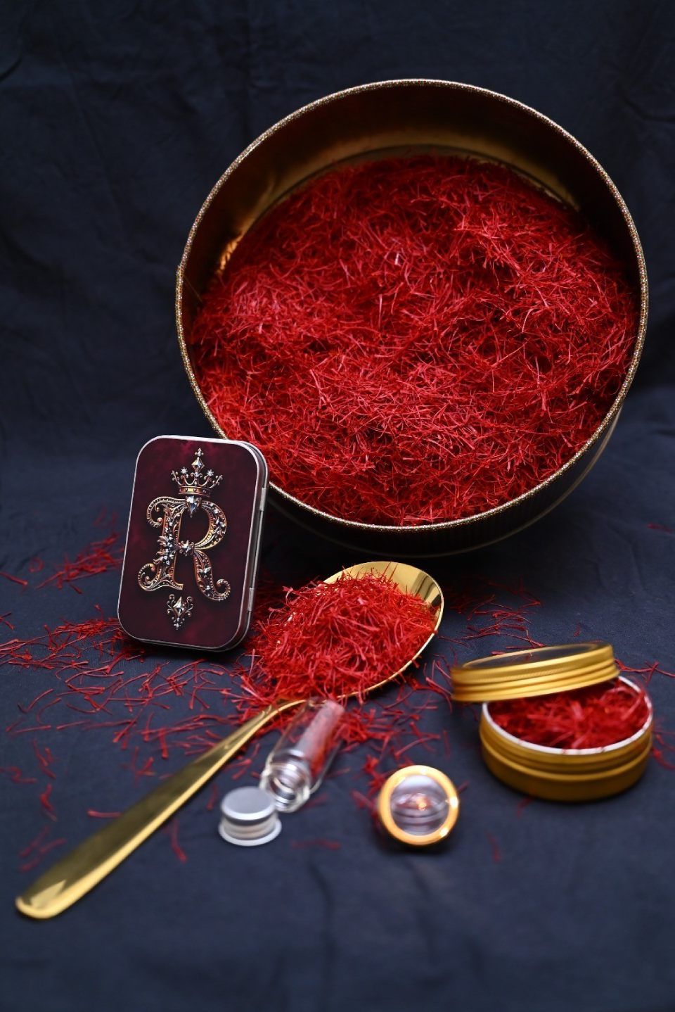 Eine Metalldose, ein Löffel und ein kleines Fläschchen gefüllt mit leuchtend roten Safranfäden vor einem dunkelblauen Hintergrund.