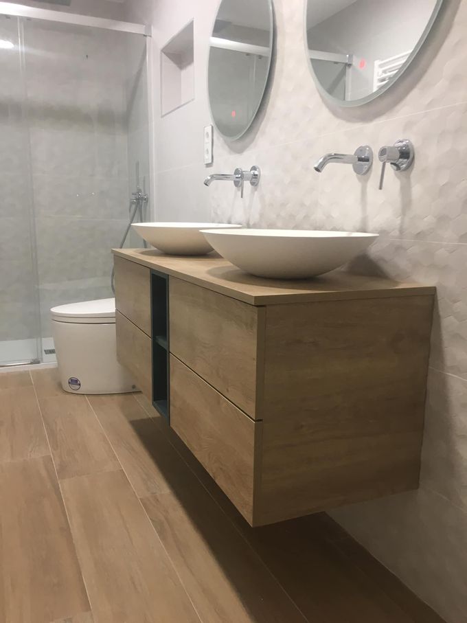 Baño moderno con tocador de madera suspendido, dos lavabos blancos sobre encimera, espejos redondos y ducha alicatada.