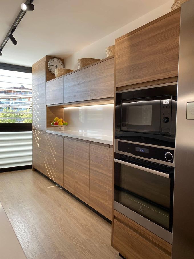 Cocina moderna con armarios de madera, horno empotrado negro, salpicadero blanco y persianas en las ventanas a la izquierda.