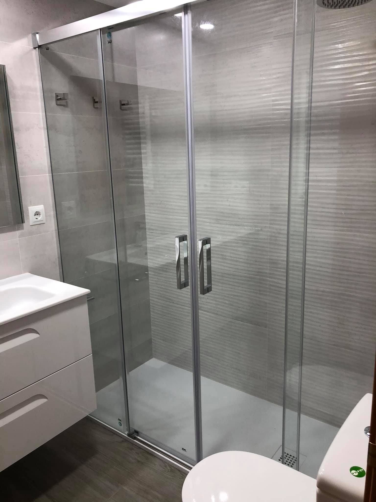 Baño moderno con cabina de ducha de cristal, paredes de azulejos grises, lavabo con mueble y inodoro.