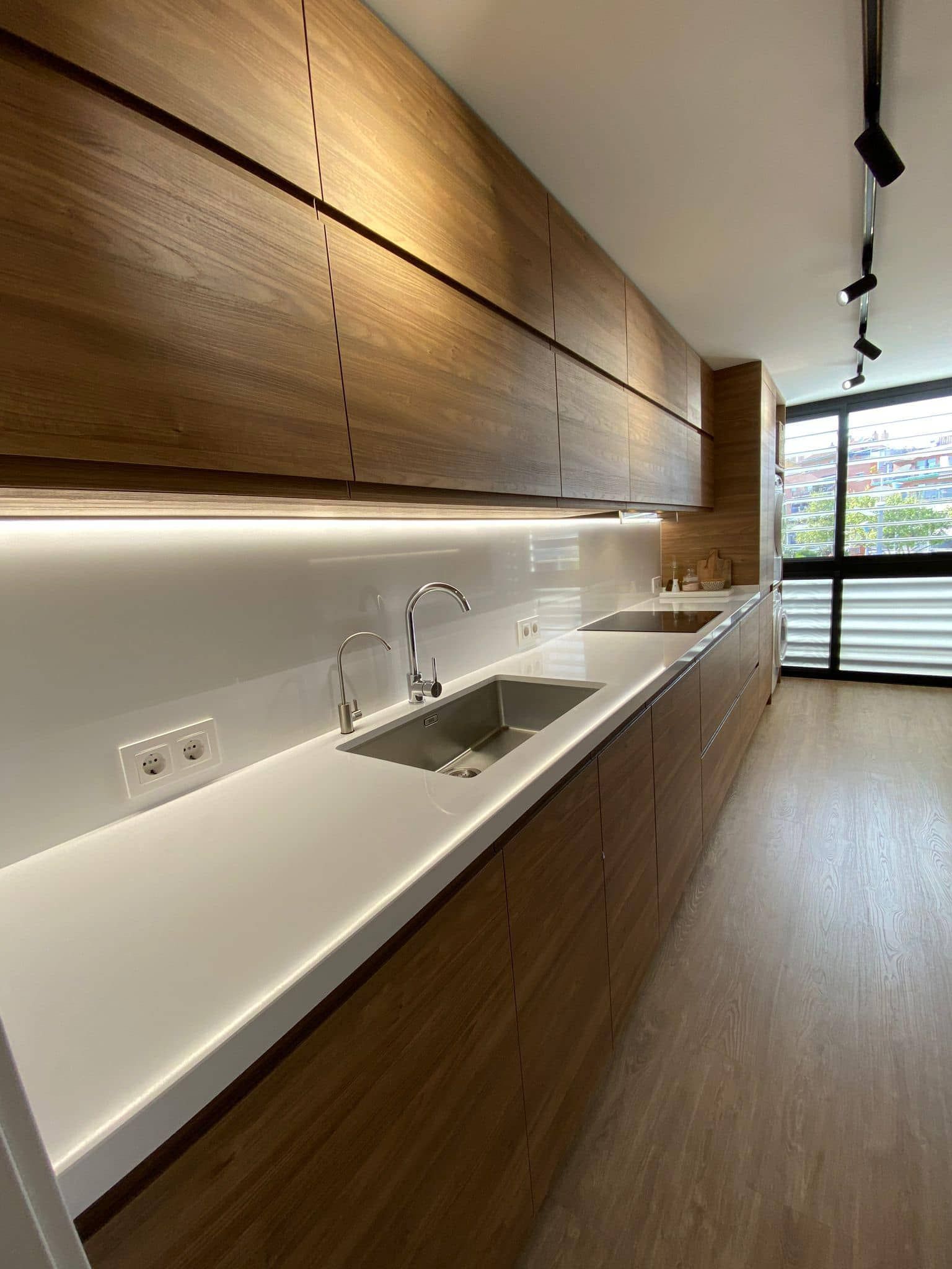 Cocina moderna tipo galera con gabinetes de madera, encimeras blancas, fregadero y grandes ventanas a lo largo de una pared.