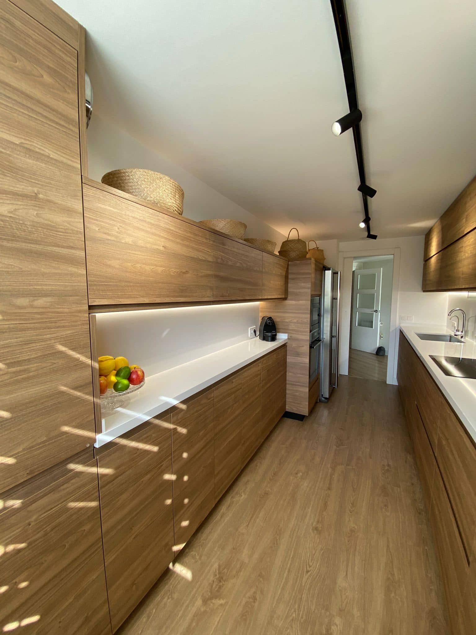Cocina moderna y estrecha con armarios de madera, encimeras blancas y vistas a un pasillo que da a una puerta.