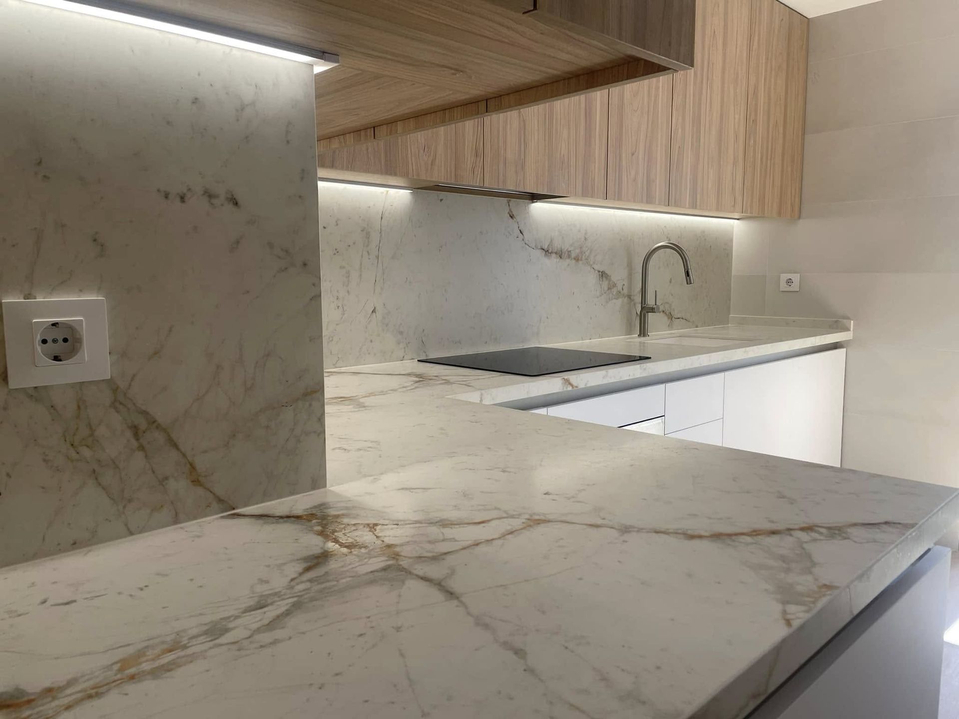 Rincón de cocina moderno con encimeras de mármol blanco, armarios de madera clara, fregadero e iluminación bajo los armarios.