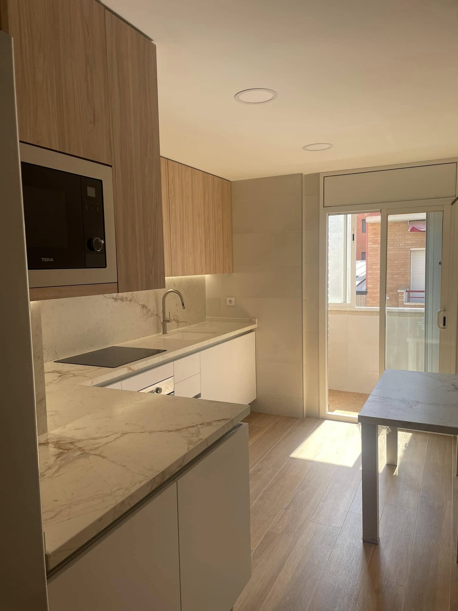 Cocina moderna con gabinetes de madera, encimeras de mármol, isla central y luz natural que entra por las puertas del balcón.