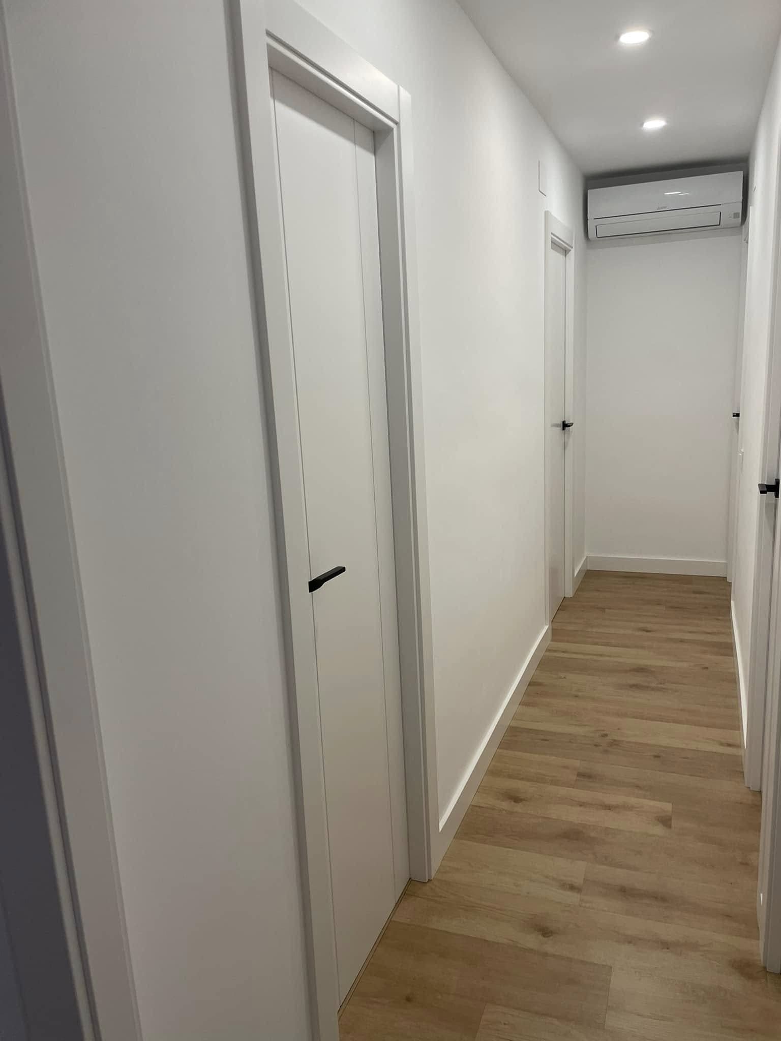 Pasillo estrecho de color blanco con suelo de madera y varias puertas cerradas a ambos lados.