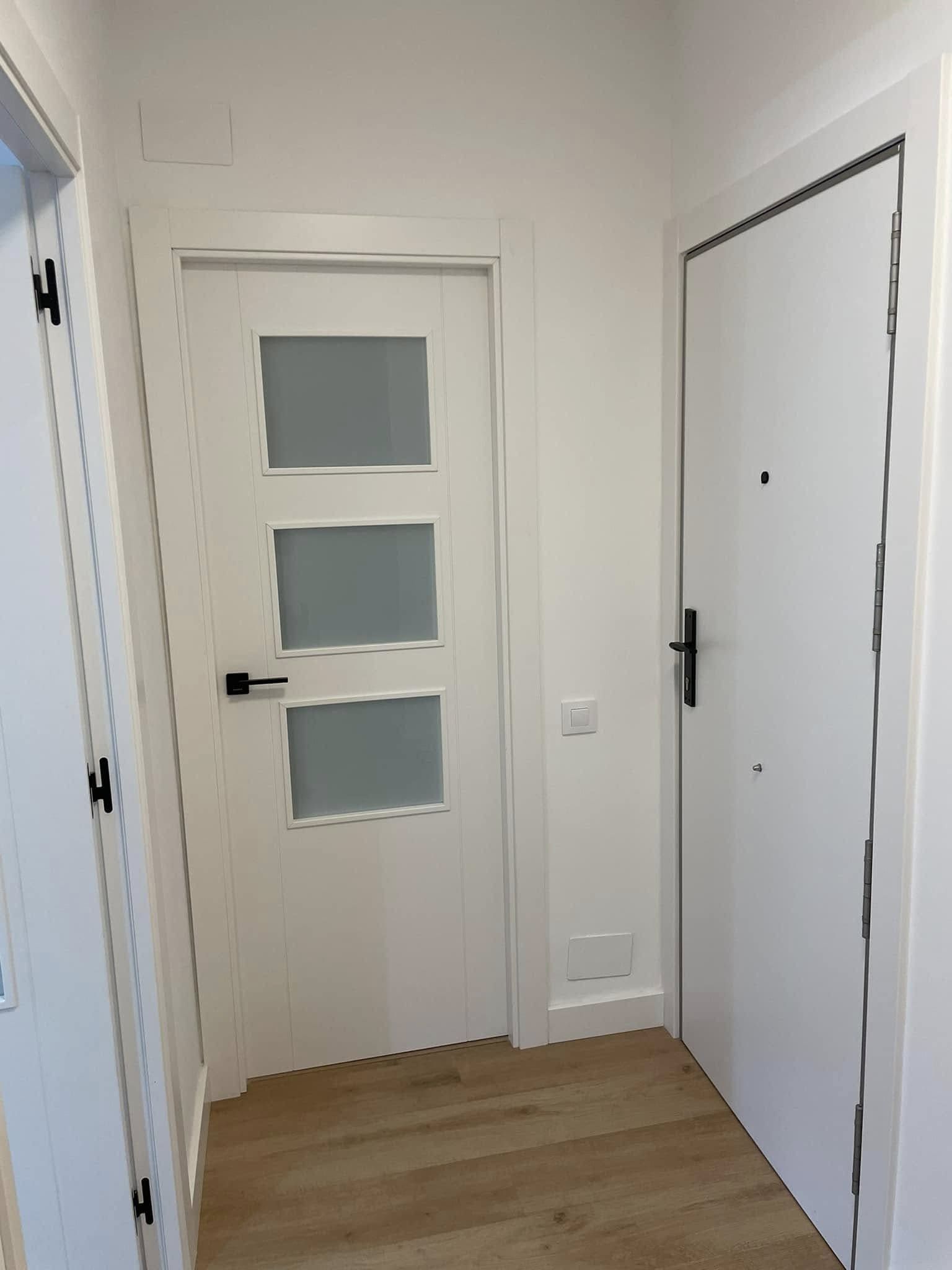 Pasillo estrecho con puertas blancas y suelo de madera clara, incluyendo una puerta con tres paneles de vidrio esmerilado.