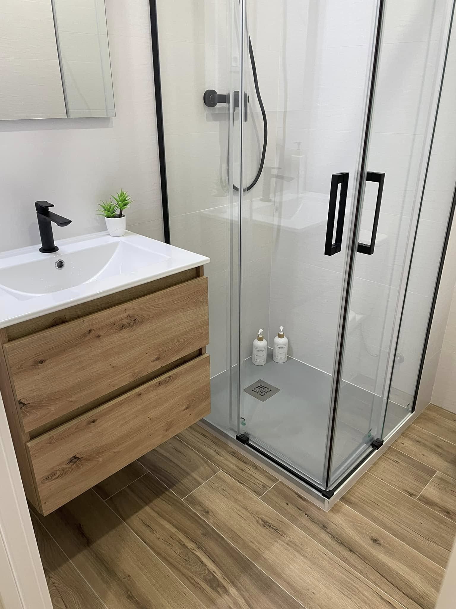 Baño moderno con lavabo blanco, tocador de madera, ducha de cristal y suelo de madera clara.