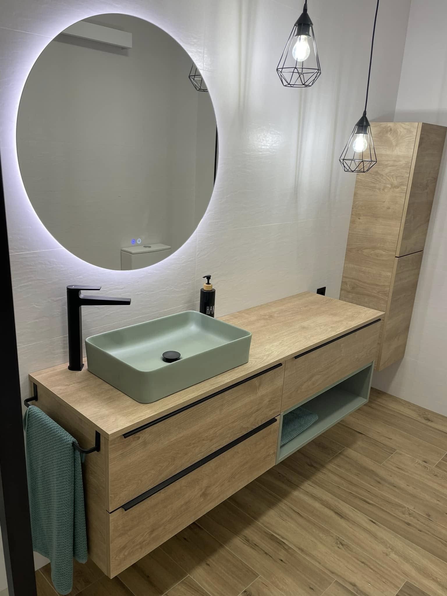 Tocador de baño moderno con espejo redondo, mueble de madera clara, grifo negro y encimera de lavabo verde.
