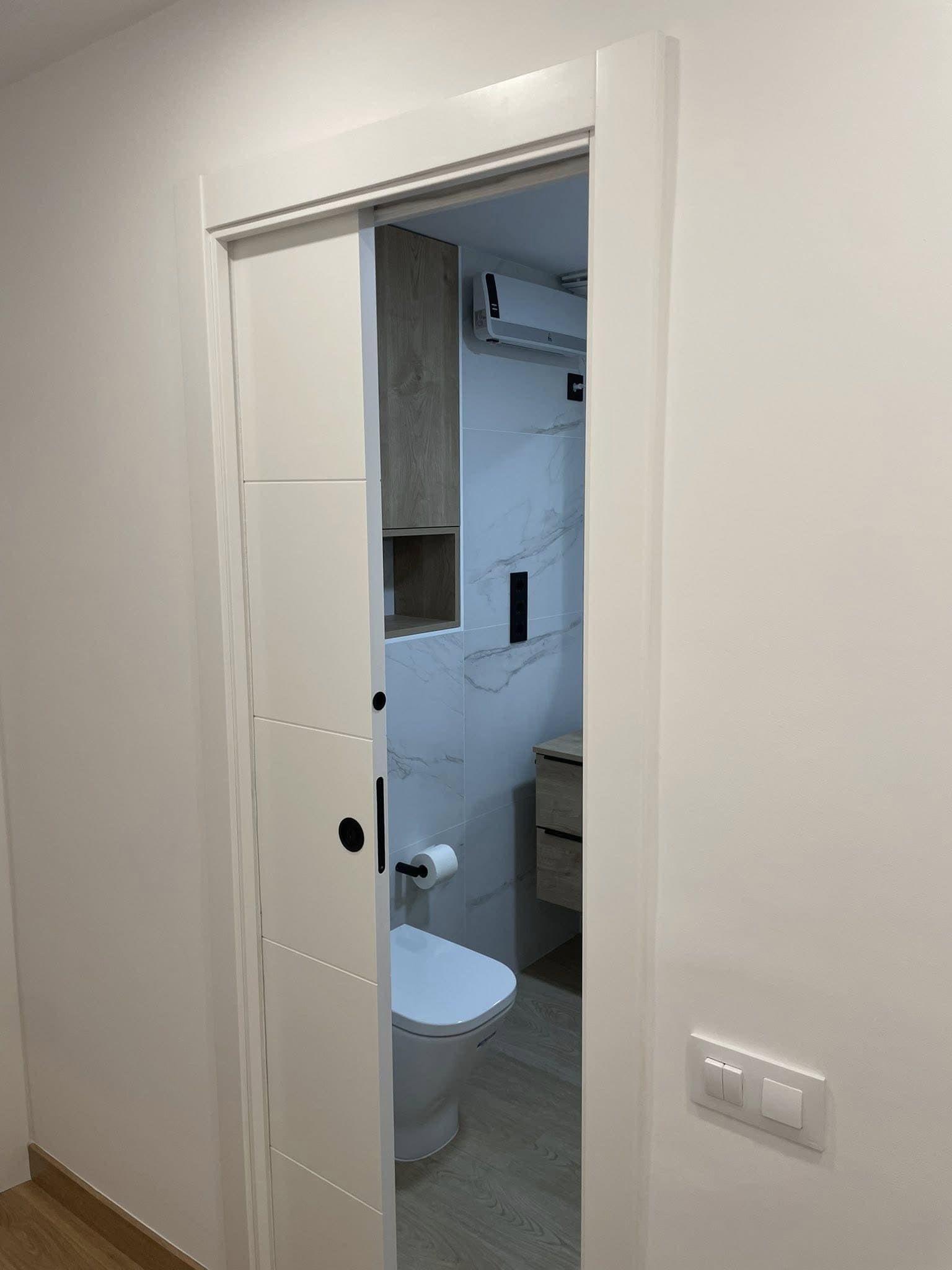 Entrada de baño con una puerta blanca entreabierta y un inodoro visible en el interior.