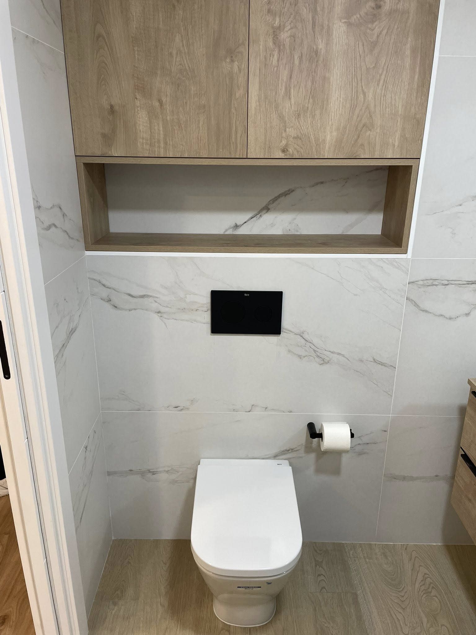 Baño moderno con inodoro suspendido, azulejos de mármol, mueble de madera y panel de descarga negro.