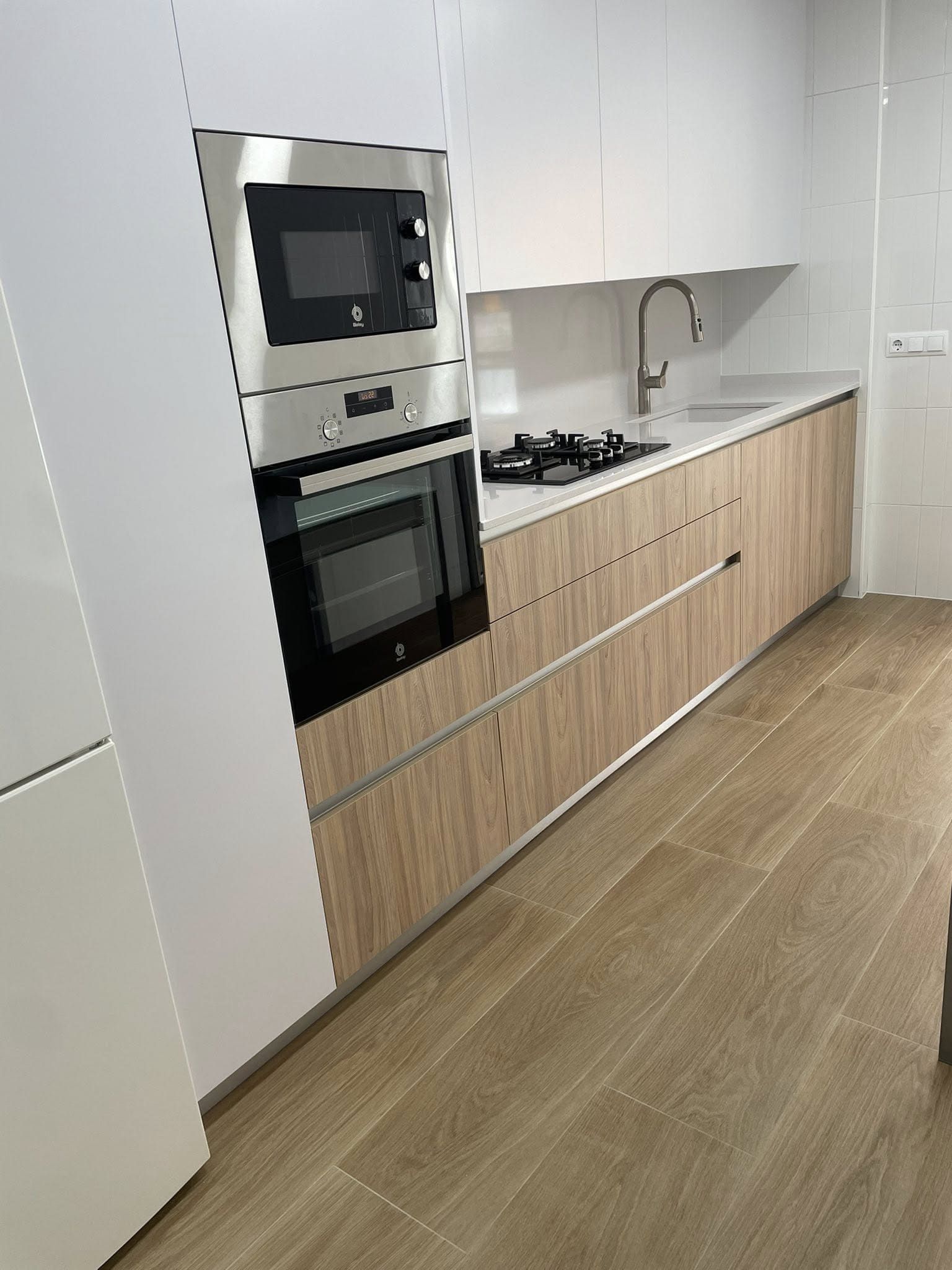Cocina moderna con horno y microondas empotrados, armarios de madera clara, salpicadero blanco y placa de gas.