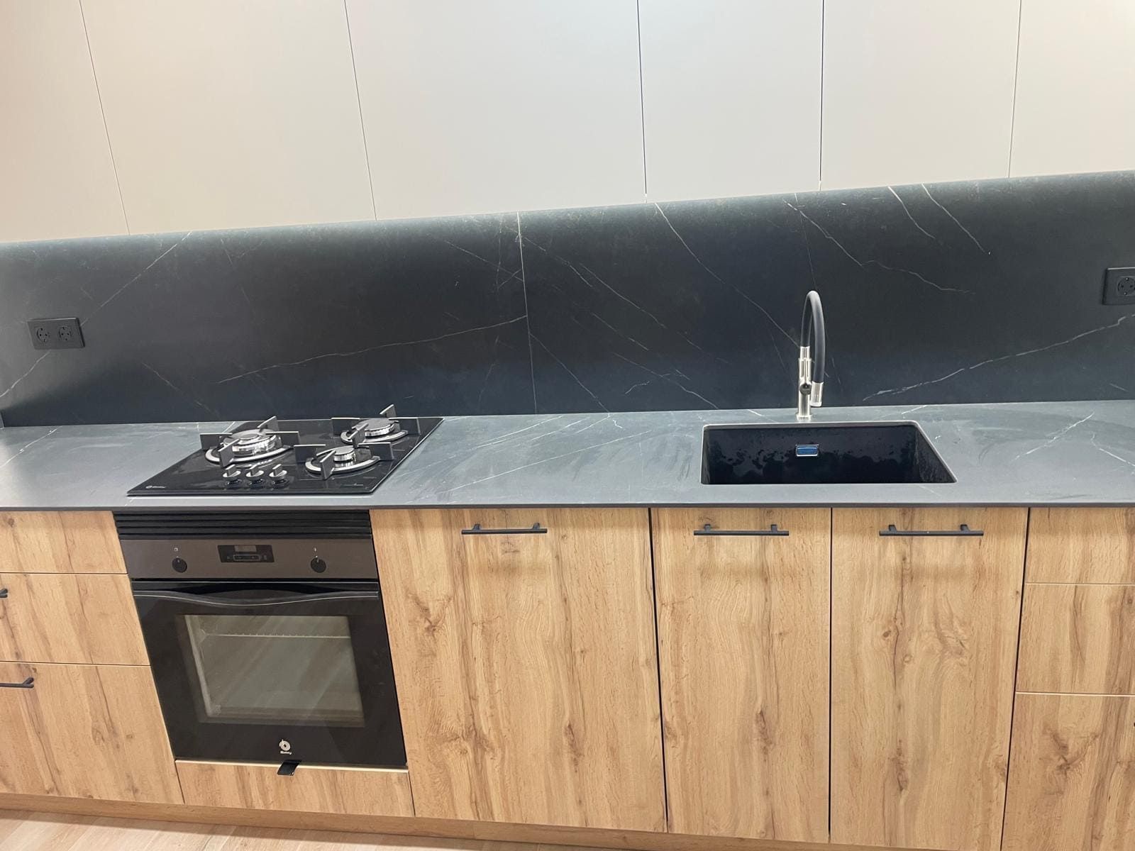 Cocina moderna con gabinetes de madera, encimera oscura, placa de gas, horno y fregadero negro sobre un salpicadero gris.