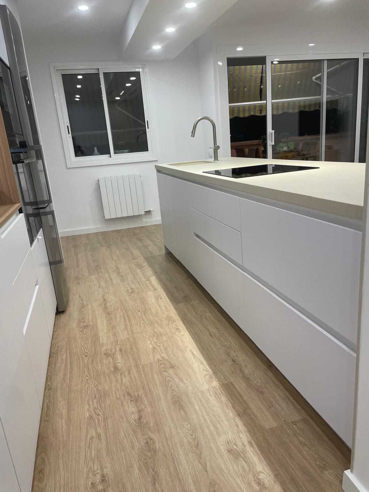 Cocina moderna blanca con isla, suelo de madera, fregadero, ventana y luces empotradas en el techo.