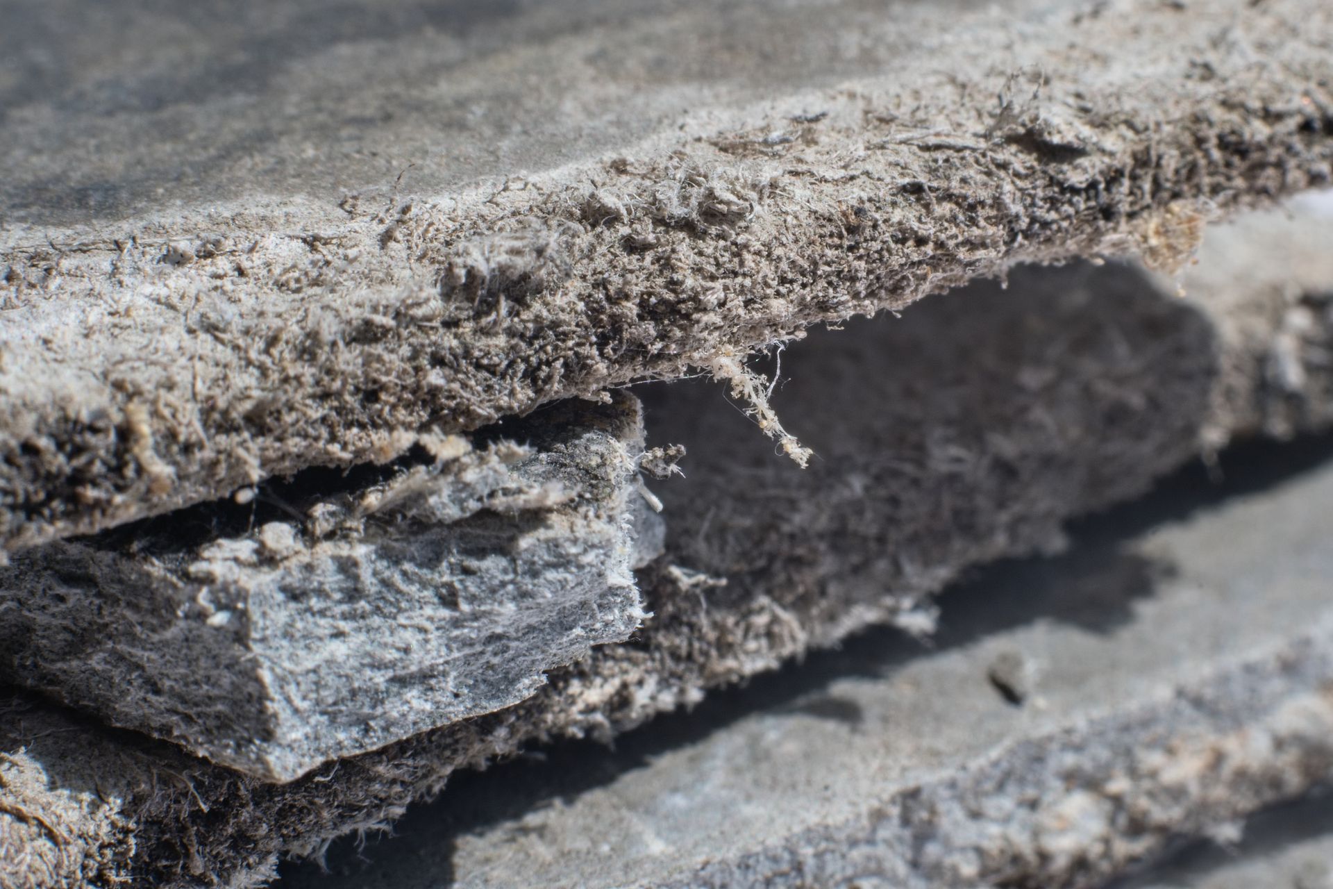 Gros plan sur une superposition de carreaux d'amiante gris altérés, avec une texture fibreuse visible et une section coupée.