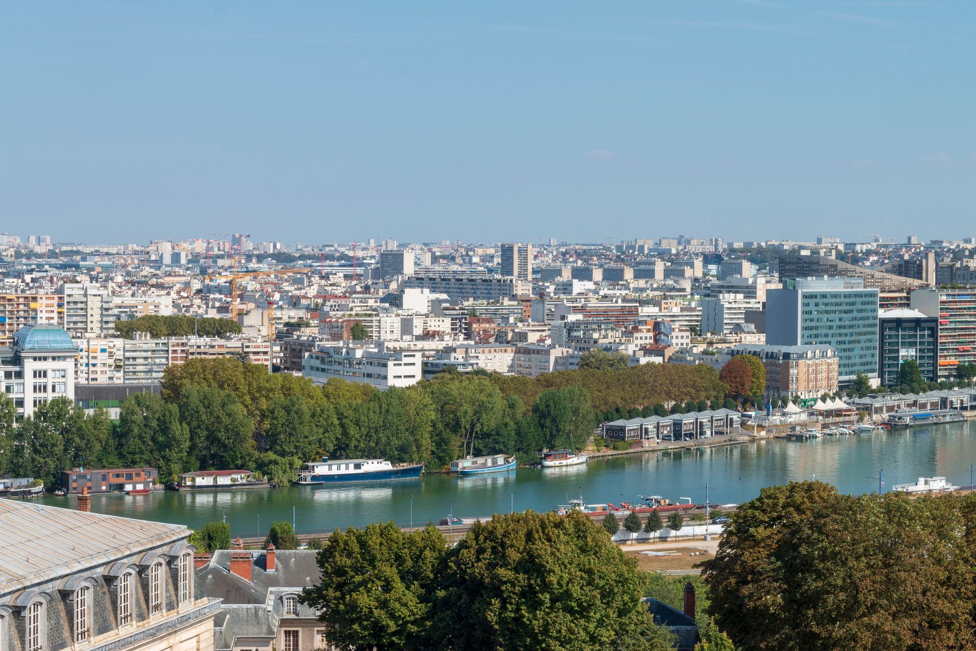 Paysage urbain avec rivière et bâtiments sous un ciel bleu ; arbres et bateaux au premier plan.