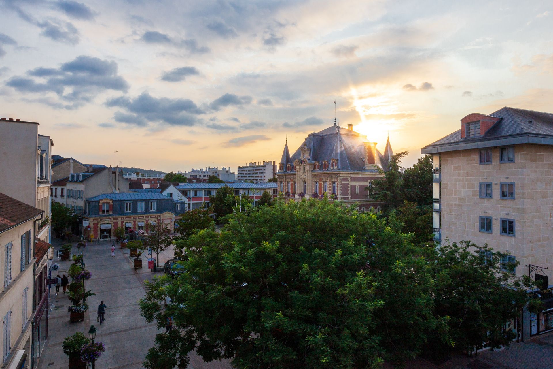 Paysage urbain au coucher du soleil, avec un bâtiment central baigné d'une lumière dorée. Des arbres et d'autres bâtiments l'entourent.