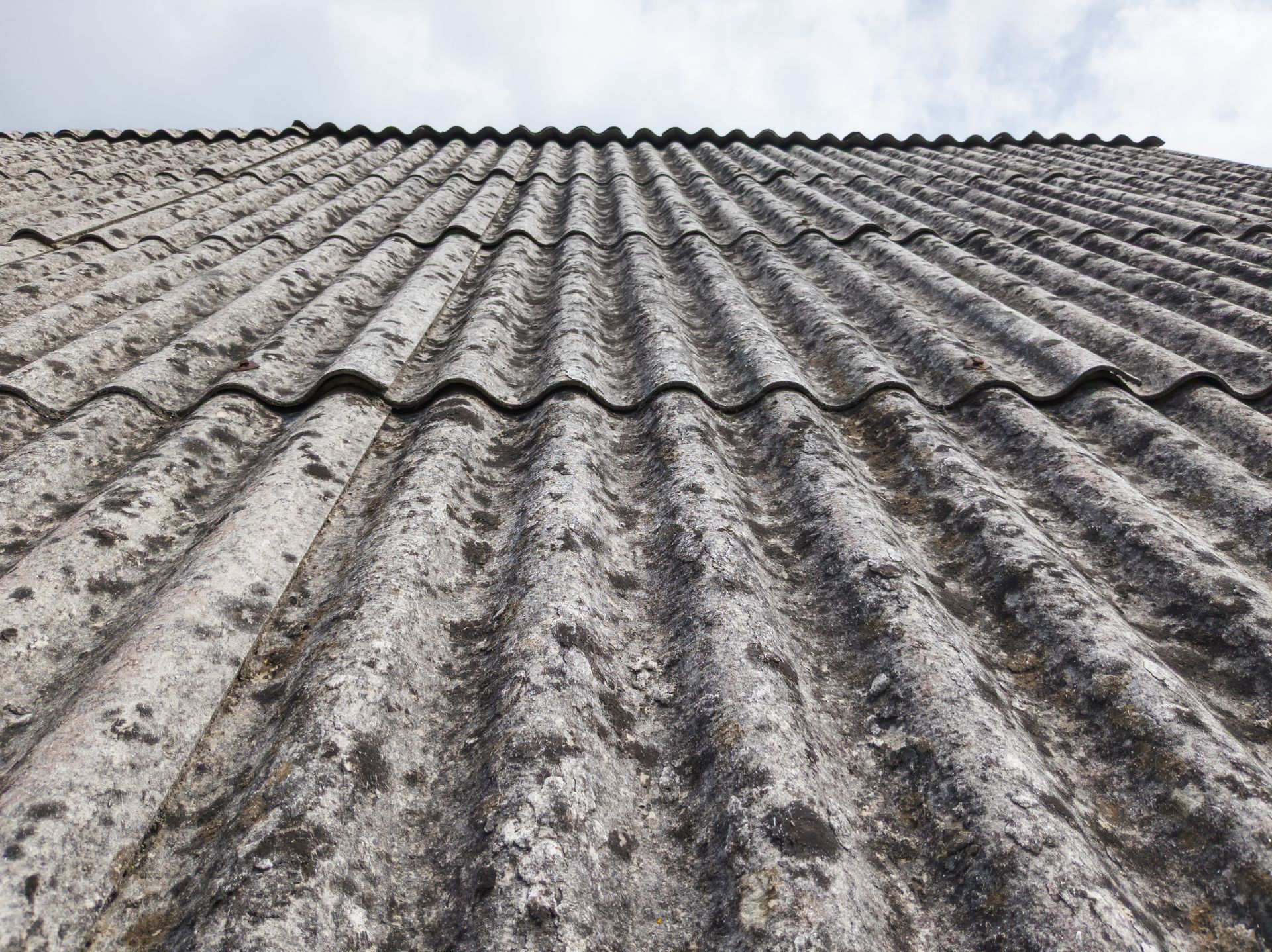 Toit en amiante grise et ondulée sous un ciel nuageux, présentant des signes d'usure et de mousse.