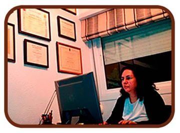 Mujer sentada en un escritorio de una oficina, mirando un ordenador debajo de unos certificados enmarcados en la pared.