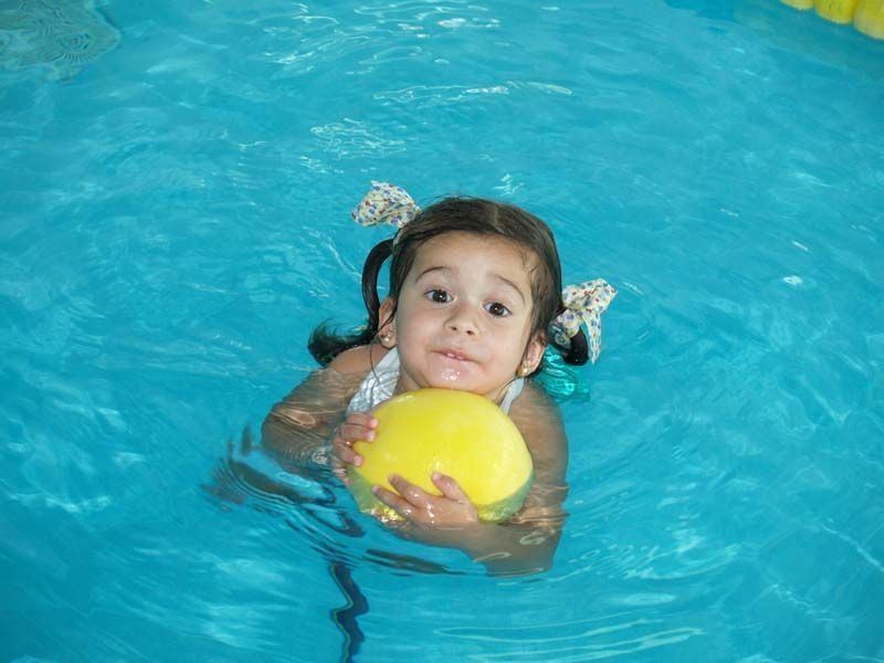 Chica en una piscina sosteniendo una pelota amarilla, sonriendo con coletas.
