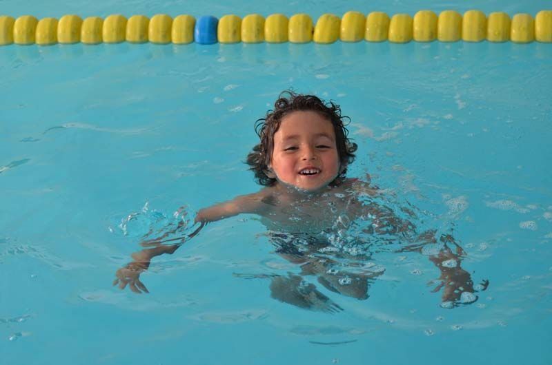 Niño pequeño con cabello rizado nadando en una piscina, sosteniendo un animal de peluche, sonriendo.