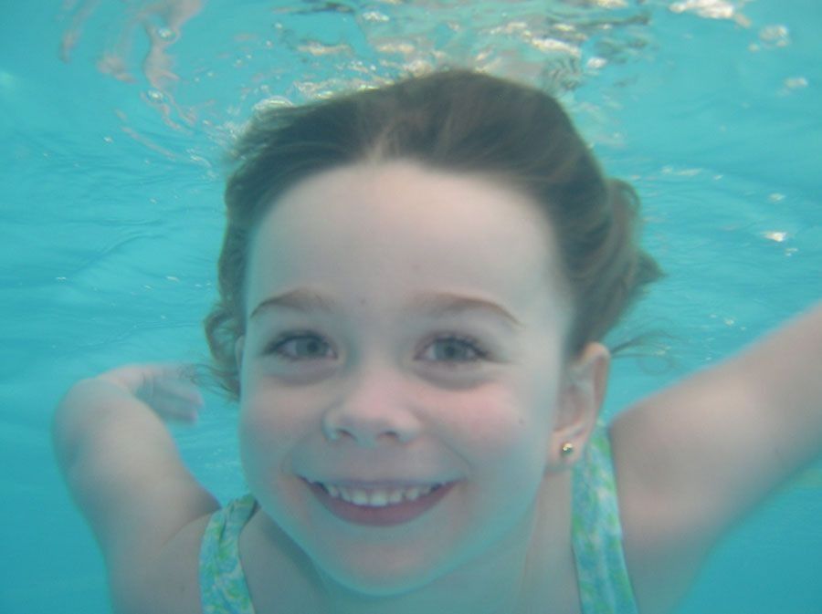 Chica sonriendo bajo el agua en una piscina, vistiendo un traje de baño.
