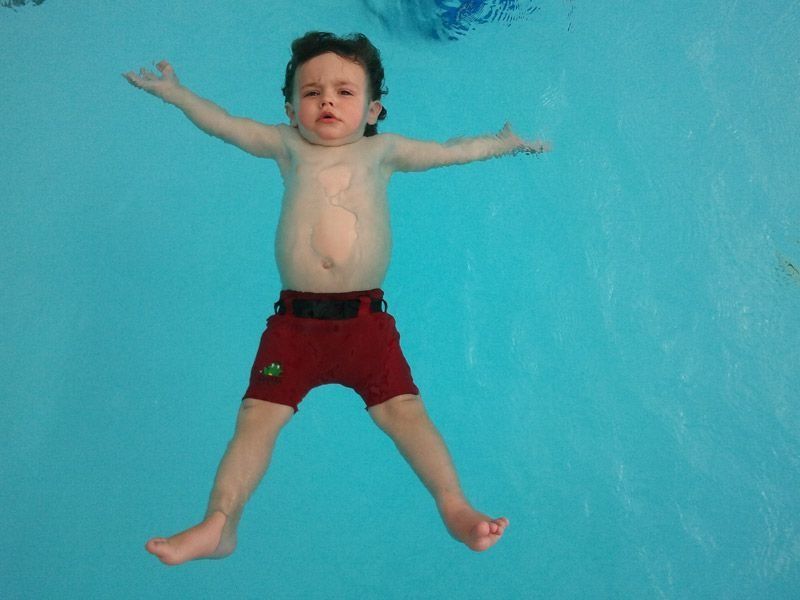 Un niño flota en una piscina, con los brazos y las piernas extendidos, vistiendo un bañador rojo, agua azul.