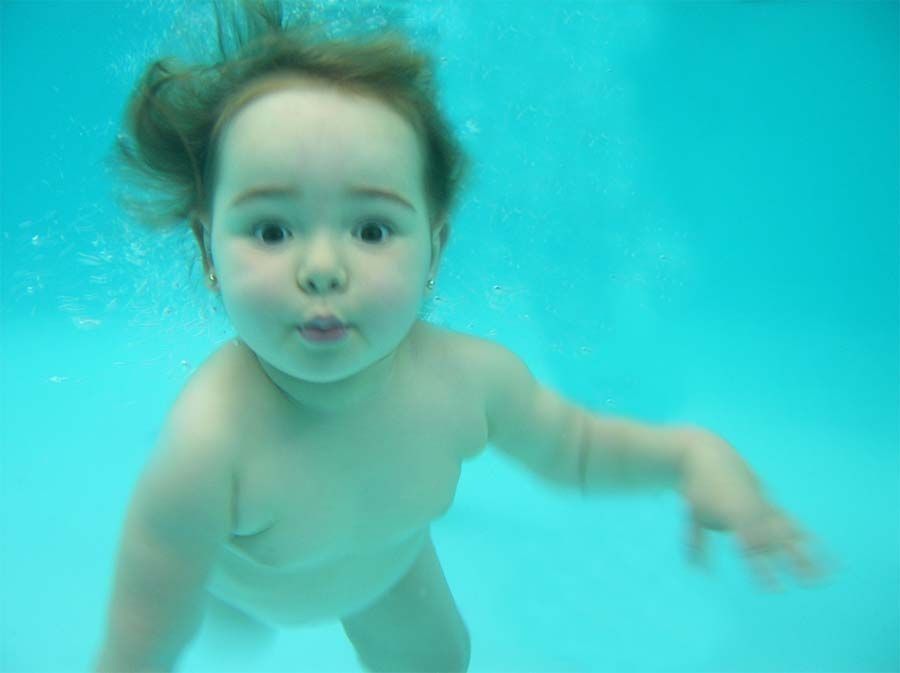 Bebé nadando bajo el agua, con los ojos muy abiertos y el cabello flotando, fondo de agua azul.