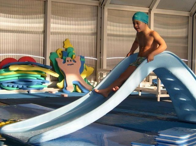 Un niño con un gorro de baño azul se desliza por un pequeño tobogán hacia una piscina.