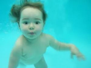 Bebé nadando bajo el agua, mirando directamente a la cámara con una expresión inquisitiva.