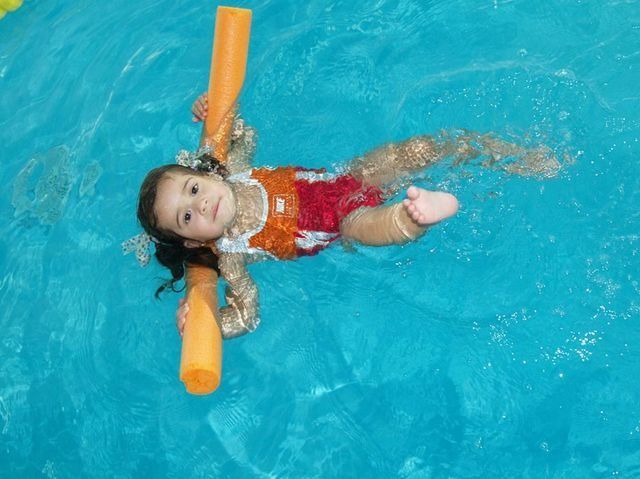 Una chica en traje de baño rojo flota en la piscina, sosteniendo fideos de piscina naranjas y mirando hacia arriba.