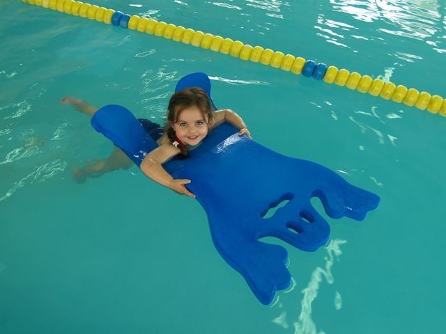 Niña sonriendo, flotando en un juguete de espuma azul en una piscina cubierta.