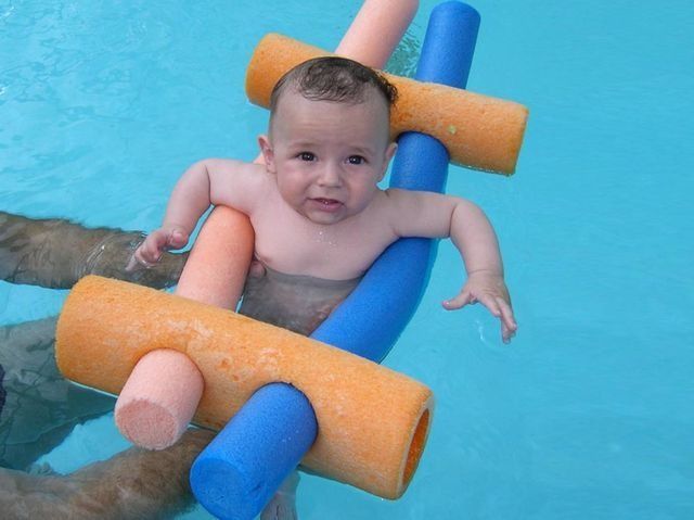 Bebé en la piscina, rodeado de flotadores de espuma naranja y azul. Mira hacia arriba con expresión neutra.
