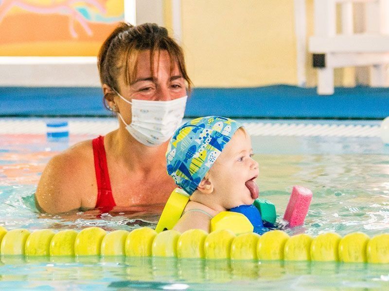 Mujer con mascarilla enseña a bebé a nadar en la piscina.