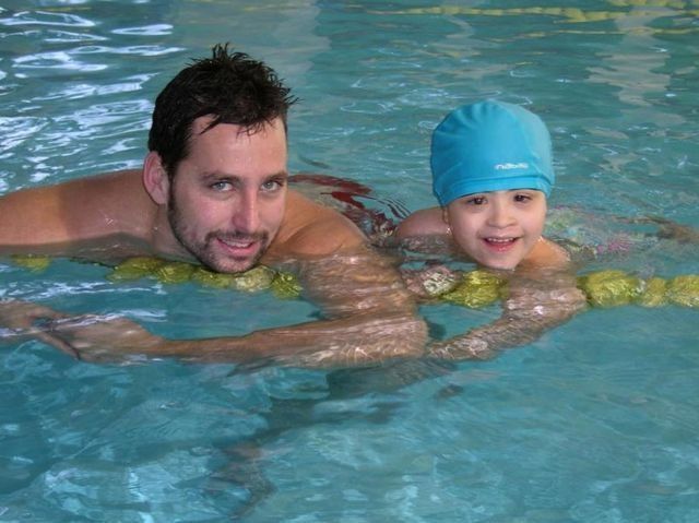 Hombre y niño en una piscina, sonriendo. El hombre no lleva camisa y el niño lleva un gorro de baño azul.