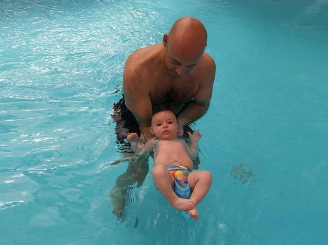 Hombre sostiene a un bebé en una piscina; el bebé lleva un pañal de baño y el hombre está sonriendo.