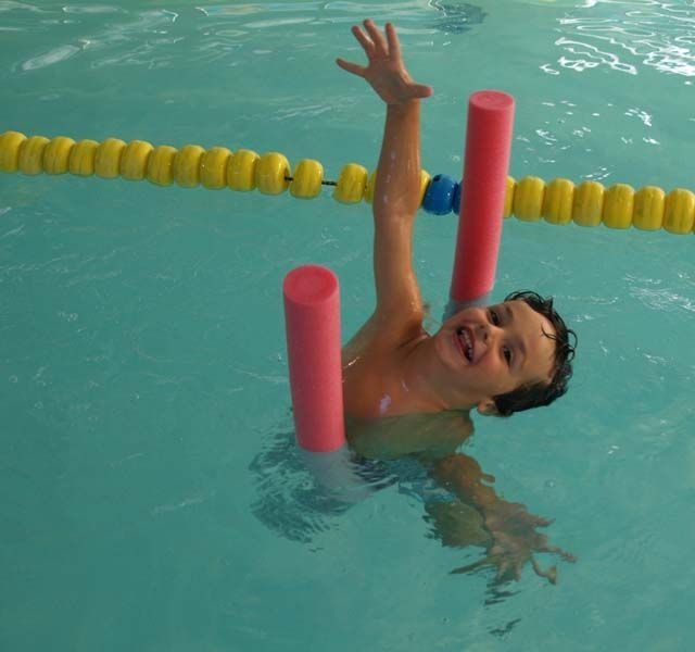 Un niño en una piscina, usando fideos de piscina rosados como apoyo, sonríe y levanta la mano.