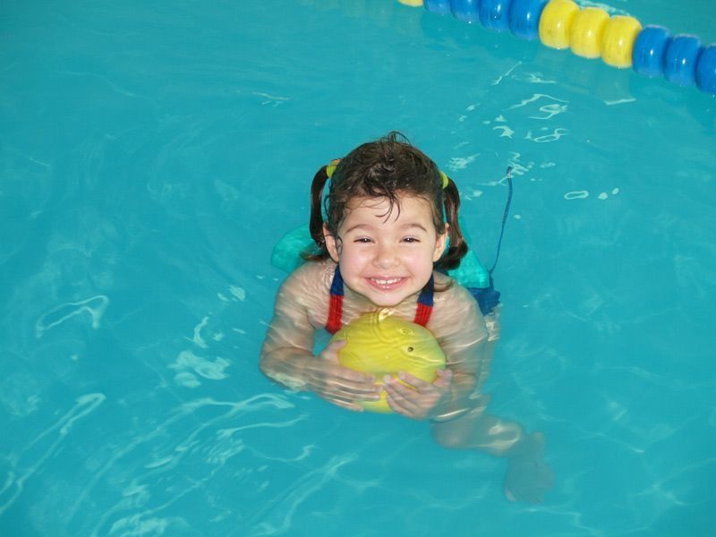 Niña en una piscina azul sonriendo, sosteniendo una pelota amarilla, usando un dispositivo de flotación, marcadores de carril azules y amarillos.