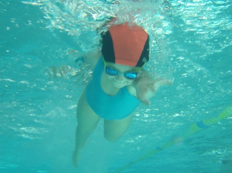 Chica bajo el agua, nadando estilo libre en traje de baño azul, gorro de baño naranja, gafas protectoras, mirando hacia la cámara.