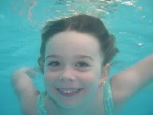 Niña sonriendo bajo el agua, con los ojos abiertos, en una piscina.